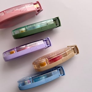 Peut inclure: Cinq distributeurs de ruban adh&eacute;sif color&eacute;s avec des corps en plastique transparent et des rubans de diff&eacute;rentes couleurs. Les distributeurs sont dispos&eacute;s en demi-cercle sur une surface blanche.