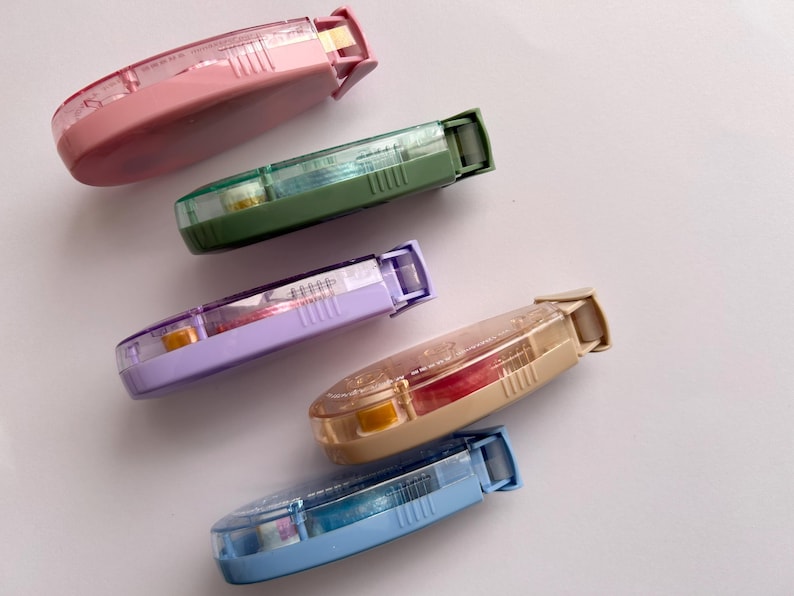 Peut inclure: Cinq distributeurs de ruban adh&eacute;sif color&eacute;s avec des corps en plastique transparent et des rubans de diff&eacute;rentes couleurs. Les distributeurs sont dispos&eacute;s en demi-cercle sur une surface blanche.