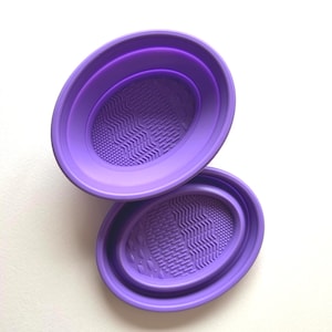 May include: Two oval-shaped, purple makeup brush cleaning mats. The mats have textured surfaces for cleaning brushes. The mats are made of a flexible material and are designed to fit inside each other for storage.