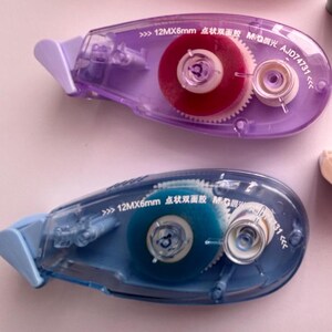 Peut inclure: Deux distributeurs de ruban adh&eacute;sif en plastique transparent avec des corps bleu et violet. Les distributeurs ont une petite fen&ecirc;tre transparente montrant le ruban &agrave; l'int&eacute;rieur. Le ruban est bleu dans un distributeur et rouge dans l'autre. Les distributeurs ont du texte sur le c&ocirc;t&eacute; qui dit "12MX6mm 点状双面胶 MG限光 AJD74731".