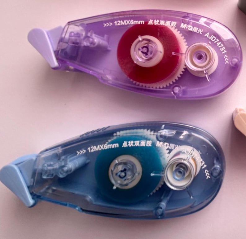 Peut inclure: Deux distributeurs de ruban adh&eacute;sif en plastique transparent avec des corps bleu et violet. Les distributeurs ont une petite fen&ecirc;tre transparente montrant le ruban &agrave; l'int&eacute;rieur. Le ruban est bleu dans un distributeur et rouge dans l'autre. Les distributeurs ont du texte sur le c&ocirc;t&eacute; qui dit "12MX6mm 点状双面胶 MG限光 AJD74731".