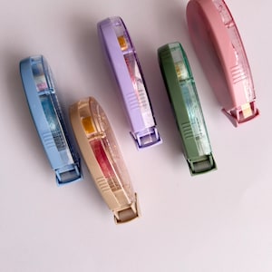 Peut inclure: Cinq distributeurs de ruban adh&eacute;sif color&eacute;s avec des corps en plastique transparent et des rubans de diff&eacute;rentes couleurs. Les distributeurs sont dispos&eacute;s en rang&eacute;e sur une surface blanche.
