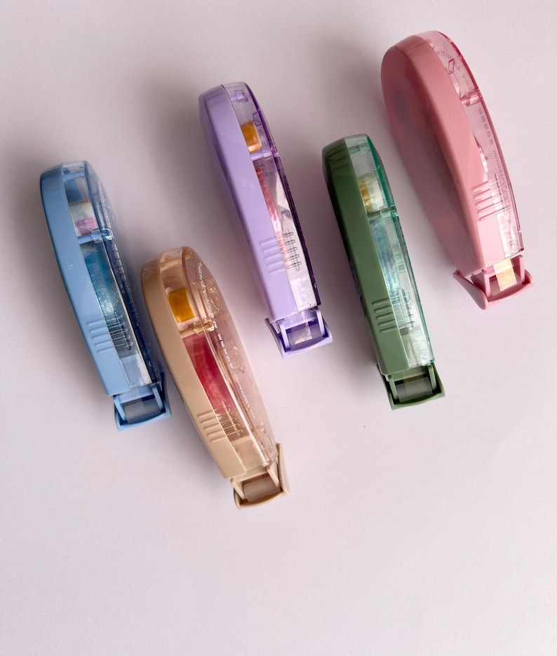 Peut inclure: Cinq distributeurs de ruban adh&eacute;sif color&eacute;s avec des corps en plastique transparent et des rubans de diff&eacute;rentes couleurs. Les distributeurs sont dispos&eacute;s en rang&eacute;e sur une surface blanche.