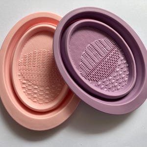 Könnte beinhalten: Zwei Silikon-Reinigungspads für Make-up-Pinsel. Einer ist rosa und der andere lila. Beide haben texturierte Oberflächen zum Reinigen von Make-up-Pinseln.