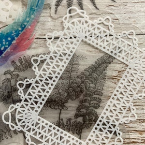 May include: White decorative frame with a delicate lace design. The frame is made of a thin, translucent material and has a square shape. The frame is perfect for adding a touch of elegance to any project.