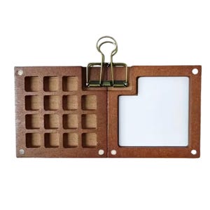 Pode incluir: Um organizador de mesa de madeira com um design articulado. Um lado apresenta 16 pequenos compartimentos quadrados, enquanto o outro tem um espaço retangular branco. Uma presilha de metal prende os dois lados. O organizador é feito de madeira marrom.