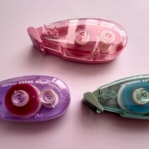 Peut inclure: Trois distributeurs de ruban adh&eacute;sif transparent avec des rouleaux de ruban de couleurs diff&eacute;rentes. Les distributeurs sont rose, violet et bleu. Les rouleaux de ruban sont rose, rouge et bleu. Les distributeurs sont &eacute;tiquet&eacute;s avec le texte "5,5 x 12 mm x 6 mm  CORRECTION TAPE  AJ014731  6,5 m".