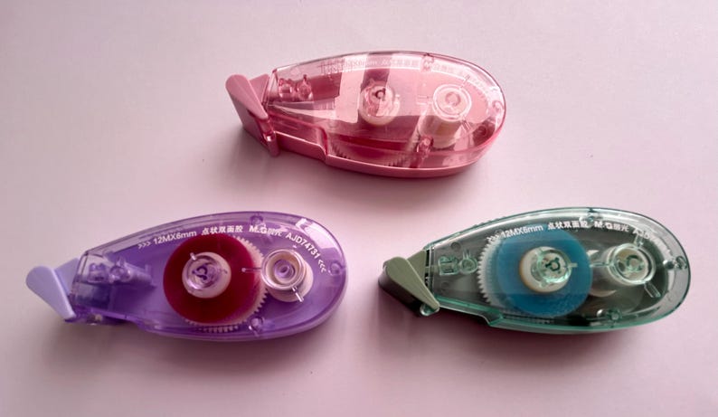 Peut inclure: Trois distributeurs de ruban adh&eacute;sif transparent avec des rouleaux de ruban de couleurs diff&eacute;rentes. Les distributeurs sont rose, violet et bleu. Les rouleaux de ruban sont rose, rouge et bleu. Les distributeurs sont &eacute;tiquet&eacute;s avec le texte "5,5 x 12 mm x 6 mm  CORRECTION TAPE  AJ014731  6,5 m".