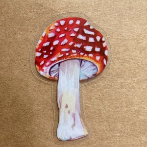 May include: A red and white mushroom sticker with a white stem. The mushroom has a rounded cap with white spots on a red background.