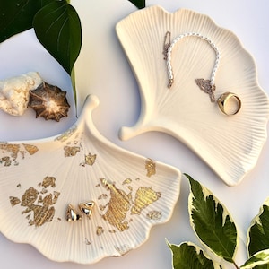 Könnte beinhalten: Zwei cremefarbene Keramiktabletts in Form von Ginkgoblättern mit Goldblattakzenten. Eines enthält goldene Ohrringe, das andere einen goldenen Ring und eine weiße Perlenkette. Grüne Blätter und Muscheln sind ebenfalls vorhanden.
