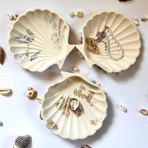 Könnte beinhalten: Drei cremefarbene, muschelförmige Schmuckschalen mit Gold- und Silberblatt-Akzenten. Eine Schale enthält einen goldenen Ring, eine andere eine Halskette und ein Armband. Kleine Muscheln sind verstreut.
