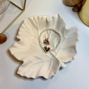 Könnte beinhalten: Eine weiße, blattförmige Keramikschale enthält ein Armband mit Perlen und ein Paar goldfarbene herzförmige Ohrringe. Die Ohrringe haben eine polierte Oberfläche. Das Armband besteht aus kleinen, weißen Perlen.