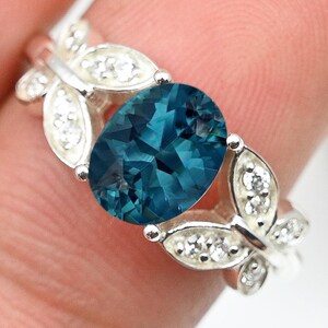 May include: A silver ring featuring an oval, teal-colored gemstone as the centerpiece. The ring's band is adorned with leaf-shaped designs, each accented with small, clear gemstones. The ring is held between fingers.