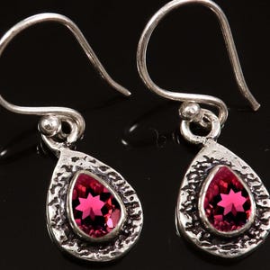 Może przedstawiać: Para srebrnych kolczyków wiszących. Każdy kolczyk ma teksturowaną oprawę w kształcie łzy z żywym, fasetowanym, magentowym kamieniem szlachetnym. Kolczyki mają prosty haczykowy design do przekłutych uszu.