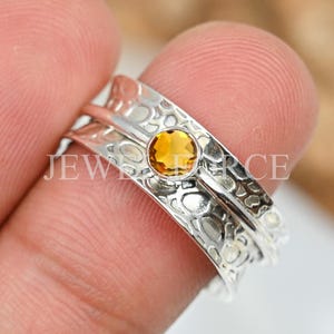 May include: A silver spinner ring featuring a textured, hammered surface and a single, round, faceted yellow citrine gemstone set in a smooth bezel. The band has a wide, flat profile with a polished finish on the edges and a recessed center.