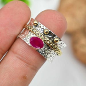 Op de afbeelding: Een zilveren ring met een robijn edelsteen. De ring heeft een textuurontwerp met gouden accenten en kleine gouden kralen. De robijn is ovaal van vorm en heeft een dieprode kleur. De ring wordt vastgehouden door de vingers van een persoon.
