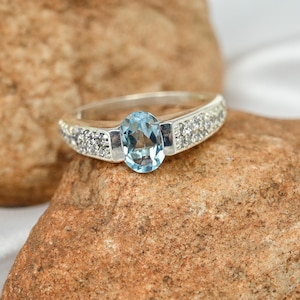 May include: A silver ring with an oval, light blue gemstone as the centerpiece. The band is adorned with small, clear gemstones on either side of the main stone. The ring is set against a textured, brown background.