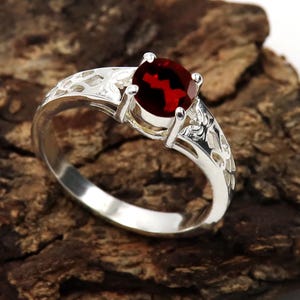 May include: A silver ring with an oval, deep red gemstone. The ring has an ornate, openwork design on the band. The gemstone is held in place by four prongs.