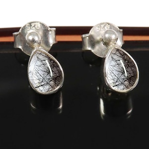May include: A pair of silver stud earrings. Each earring has a teardrop-shaped, clear stone with black inclusions, set in a silver bezel. The earrings have a post and butterfly backing. The metal is stamped with "925".