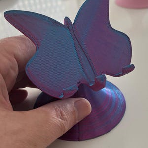 Könnte beinhalten: Ein 3D-gedruckter Handyhalter in Schmetterlingsform in Lila- und Blautönen. Der Ständer hat ein detailliertes Schmetterlingsdesign mit strukturierten Flügeln und einem kreisförmigen Sockel. Der Ständer wird von einer Hand gehalten.