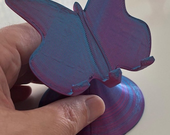 以下が含まれることがあります： 紫と青の色合いの、3Dプリントされた蝶の形をした携帯電話スタンド。スタンドは、テクスチャ加工された羽と円形のベースを備えた詳細な蝶のデザインが特徴です。スタンドは手で持たれています。