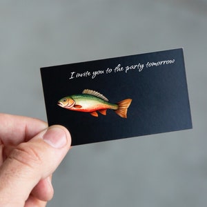 Pu&ograve; includere: Un biglietto nero con testo bianco che dice "Ti invito alla festa domani". Un'illustrazione di un pesce verde, rosso e arancione &egrave; sul biglietto.