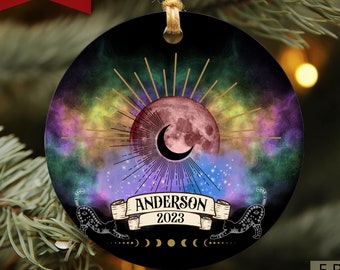 May include: A round, black, personalized Christmas ornament with a colorful nebula background. The ornament features a gold sun with a pink moon and a crescent moon. The text "Anderson 2023" is on a banner with a black cat on each side.