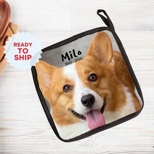 May include: A black and white pot holder with a photo of a corgi dog. The dog is looking at the camera with its tongue sticking out. The text "Milo 2017-2024" is printed on the pot holder.