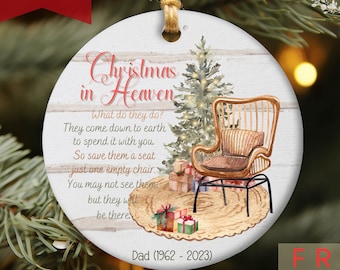 May include: A white round Christmas ornament with a watercolor illustration of a chair, a Christmas tree, and presents. The ornament has the text "Christmas in Heaven" and a quote about remembering loved ones who have passed away. The ornament also includes the text "Dad (1962 - 2023)".