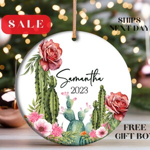 May include: A white round ceramic ornament with a watercolor design of cacti, roses, and other flowers. The ornament has the name "Samantha" and the year "2023" written on it.
