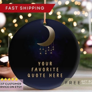 May include: Round, navy blue ornament with a gold crescent moon and hanging stars design. The text "YOUR FAVORITE QUOTE HERE" is in gold. Includes a gold string for hanging. The background is a blurred image of Christmas lights.