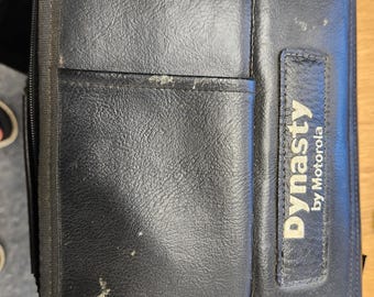 May include: Black leather case with a zippered closure and a front pocket. The case has the text "Dynasty by Motorola" printed on a rectangular patch. The case appears to be well-used.