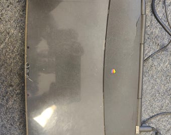 May include: A dark gray vintage laptop computer with a closed lid featuring a small, colorful Apple logo. The device has a rectangular shape with visible surface wear and a power cable connected to the side, showing a classic design aesthetic.