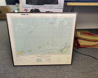 May include: Framed nautical chart of the Banda Sea, featuring detailed coastlines, navigational markers, and grid lines. The chart is set within a dark brown frame, resting on a gray carpeted surface. The map's colors include shades of blue, green, and beige.