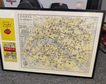 May include: Framed vintage map of Paris, France, with a detailed street layout and landmarks. The map is in a black frame and includes two yellow and red advertisements. The map is in shades of yellow, beige, and brown.