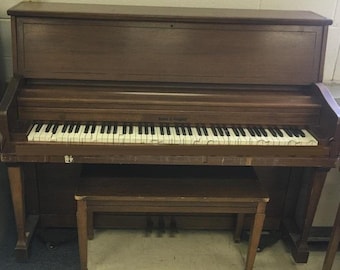 May include: A vintage upright piano with a dark wood finish. The piano has a full keyboard with black and white keys, and a matching wooden bench is positioned in front. The piano's brand name is visible.