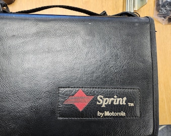 May include: Black leather case with a handle. The case features a rectangular patch with the Sprint logo in red and the text "Sprint by Motorola" in white. The case appears to be a vintage item.