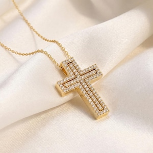 May include: A gold-colored cross pendant necklace with a delicate chain. The cross is encrusted with small, sparkling clear stones. The necklace rests on a white, silky fabric, creating a luxurious and elegant aesthetic.
