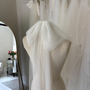 Puede incluir: Un velo de novia blanco y transparente drapeado sobre un maniquí. Otros velos blancos cuelgan en el fondo. La escena tiene lugar en una tienda o estudio de novias bien iluminado.