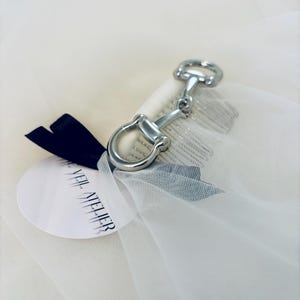 May include: A silver-colored metal horse bit keychain attached to a white veil. A black ribbon and a white tag with the text "VEIL ATELIER" are also present. The veil is made of a sheer, lightweight fabric.