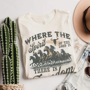 K&ouml;nnte beinhalten: Ein beiges T-Shirt mit einem Vintage-Grafikdruck von rennenden Pferden. Der Text auf dem Shirt lautet "Where the Spirit of the Lord is There is Freedom".