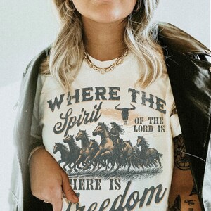 K&ouml;nnte beinhalten: Wei&szlig;es T-Shirt mit einer Grafik einer Gruppe von Pferden, die rennen. Der Text "Where the Spirit of the Lord is there is Freedom" ist auf das T-Shirt gedruckt.