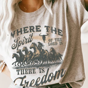 K&ouml;nnte beinhalten: Ein graues T-Shirt mit einem Vintage-Grafikdruck einer galoppierenden Pferdeherde. Der Text auf dem Shirt lautet "Where the Spirit of the Lord is There is Freedom".