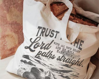 May include: A white canvas tote bag with a black and white graphic design. The graphic features a cowboy hat, a landscape, and the text "Trust in the Lord with all your heart and he will make your paths straight."
