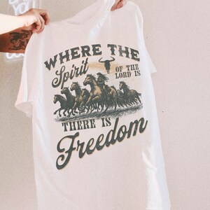 K&ouml;nnte beinhalten: Wei&szlig;es T-Shirt mit einem grafischen Druck einer Herde galoppierender Pferde mit dem Text "Where the Spirit of the Lord is There is Freedom".