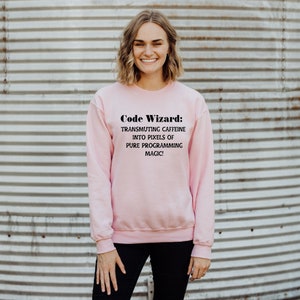 May include: A pink sweatshirt with the text "Code Wizard: Transmuting caffeine into pixels of pure programming magic!" printed on the front.