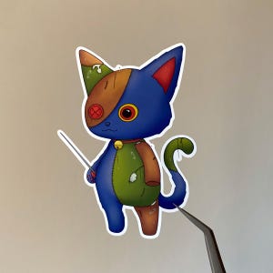 Könnte beinhalten: Ein Aufkleber mit einer Cartoon-Katze mit Patchwork-Design. Die Katze ist blau, grün und braun mit einem roten Knopfauge und einer kleinen Glocke. Sie hält Nadel und Faden und hat ein skurriles, genähtes Aussehen.
