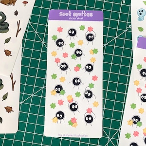 May include: A sticker sheet with cartoon soot sprites, black with white eyes and small limbs. The stickers are accompanied by colourful star-shaped stickers. The text "soot sprites" and "sticker sheet" are at the top of the sheet.