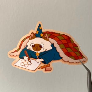 Puede incluir: Una pegatina caprichosa que presenta un gato vestido de mago, con una t&uacute;nica azul y un sombrero estrellado. El gato est&aacute; dibujando una flor en un papel, con l&aacute;pices y una goma de borrar cerca. El gato est&aacute; bajo una manta con un dise&ntilde;o de cuadros rojos y verdes.