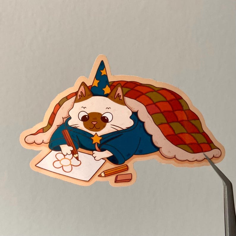 Puede incluir: Una pegatina caprichosa que presenta un gato vestido de mago, con una t&uacute;nica azul y un sombrero estrellado. El gato est&aacute; dibujando una flor en un papel, con l&aacute;pices y una goma de borrar cerca. El gato est&aacute; bajo una manta con un dise&ntilde;o de cuadros rojos y verdes.